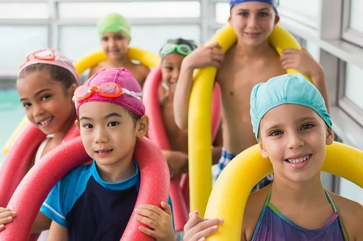 A group of kids wear swimming cap and smile with foam pool noodles around the back of their necks